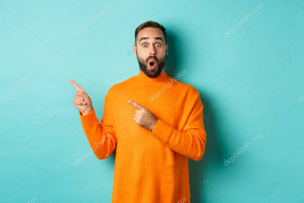 Impressed young man with beard, reacting to promo banner, pointing fingers left, showing logo and looking surprised, standing over turquoise background.