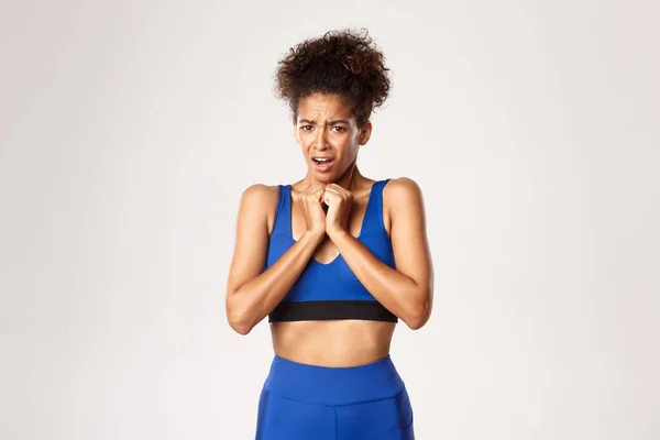 Taille-up van geschokt Afrikaans-Amerikaanse vrouwelijke atleet in blauwe sport oufit, grimmig en kijken bang, staande tegen een witte achtergrond — Stockfoto