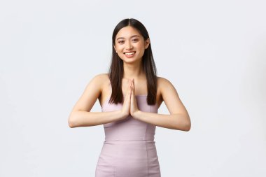 Lüks kadınlar, parti ve tatil konsepti. Akşam elbiseli çekici Asyalı kadın, el ele tutuşun ya da namaste işareti yapın, kutlama konukları selamlayın, güler yüzlü, beyaz arka plan