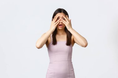 Lüks kadınlar, parti ve tatil konsepti. Korkmuş ürkek ve kendine güveni olmayan güzel Asyalı kadın gece elbisesi giymiş, korkunç duruma tanık olmuş, gözleri kapamış ve korkmuş parmaklarının arasından dikizliyormuş.