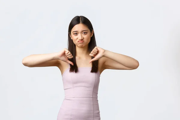 Mujeres de lujo, fiesta y concepto de vacaciones. Decepcionada mujer asiática exigente en vestido de noche, frunciendo el ceño y mueca renuente, mostrando pulgares hacia abajo en aversión, fondo blanco —  Fotos de Stock