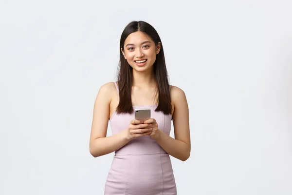 Lüks kadınlar, parti ve tatil konsepti. Gece elbisesi giymiş güzel, genç Asyalı bir kadın, cep telefonu kullanıyor, akıllı telefondan arkadaşlarıyla mesajlaşıyor, beyaz arka planda duruyor. — Stok fotoğraf