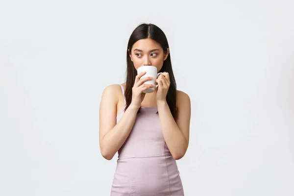 Livsstil, människors känslor och glamourkoncept. Tänksam sensuell asiatisk kvinna i aftonklänning, tittar bort tänkande och dricka kaffe från mugg, stående vit bakgrund — Stockfoto