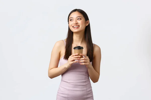 Estilo de vida, emociones de la gente y concepto de glamour. Soñadora chica asiática en vestido, sonriendo y buscando imágenes esquina superior izquierda, disfrutando de un hermoso día con taza de café, bebiendo de la taza para llevar — Foto de Stock