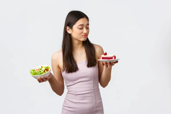 Estilo de vida saludable, ocio y concepto de comida. Hermosa chica asiática que se queda a dieta, haciendo pucheros y mirando con deseo a la torta, tentando a morder, sosteniendo ensalada otra mano, fondo blanco —  Fotos de Stock