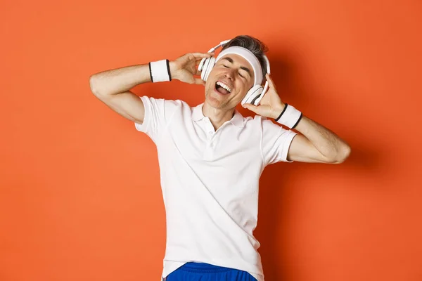 Portret beztroskiego sportowca w średnim wieku, słuchanie muzyki w słuchawkach podczas treningu fitness, stanie nad pomarańczowym tłem — Zdjęcie stockowe