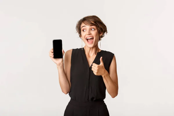 Fantastisk vacker flicka rekommenderar något på smartphone-skärmen, visar tummen upp i godkännande — Stockfoto