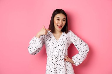 Güzel Asyalı kız mutlu görünüyor, kendini işaret ediyor, romantik pembe arka planda duruyor.