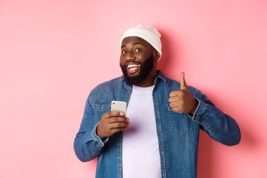 Teknoloji ve online alışveriş konsepti. Tatmin olmuş siyahi adam gülümsüyor, cep telefonu kullanıyor ve onay verirken baş parmağını kaldırıyor, düzen kuruyor, pembe arka planda duruyor.