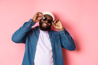 Yakışıklı Afro-Amerikalı adam güneş gözlüğü takmış, gülümsüyor memnun ve kendinden emin, pembe arka plana karşı duruyor.