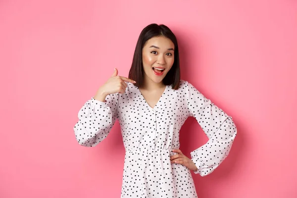 Menina asiática bonita no vestido olhando feliz, apontando o dedo para si mesma, de pé no fundo rosa romântico — Fotografia de Stock