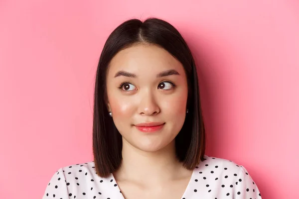 Ομορφιά και φροντίδα του δέρματος έννοια. Κοντινό πλάνο του χαριτωμένο κορίτσι της Ασίας κοιτάζοντας αριστερά στο πανό, χαμογελώντας ανόητο, στέκεται στο φόρεμα πάνω από ροζ φόντο — Φωτογραφία Αρχείου