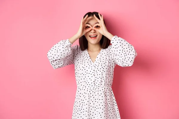 Unbekümmerte junge Asiatin blickt durch ein Fernglas in die Kamera, starrt auf Rabatte, steht über rosa Hintergrund — Stockfoto