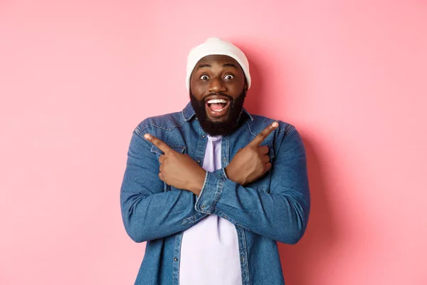 Glücklicher junger afrikanisch-amerikanischer Hipster-Typ, der die Finger zur Seite zeigt, lächelt und zwei Möglichkeiten zeigt, Angebote zeigt, vor rosa Hintergrund steht — Stockfoto
