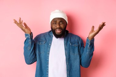 Kafası karışmış ve rahatsız olmuş siyah adam şikayet ediyor, omuz silkiyor ve ellerini kaldırıyor, hayal kırıklığıyla bakıyor, pembe arka planda duruyor.