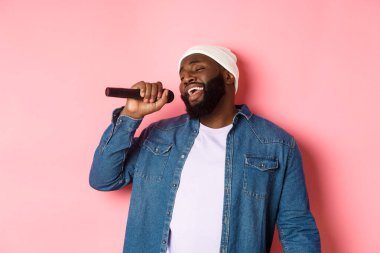 Bereli ve kot gömlekli yakışıklı siyah adam karaoke söylüyor, mikrofon tutuyor, pembe arka planda duruyor.