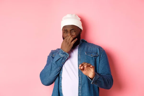 Walgelijke zwarte man sloot neus en krimp van vreselijke geur, tonen weigering gebaar, staande over roze achtergrond — Stockfoto