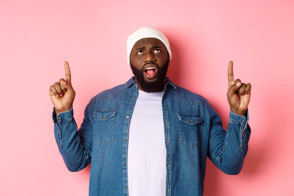 Displeased african american guy in beanie, pointing fingers up and staring at camera bothered, complaining, standing over pink background