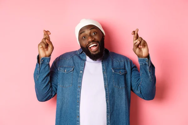 Hombre africano-americano optimista pidiendo deseo, sosteniendo los dedos cruzados y sonriendo, de pie sobre fondo rosa — Foto de Stock