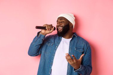 Bereli ve kot gömlekli yakışıklı siyah adam karaoke söylüyor, mikrofon tutuyor, pembe arka planda duruyor.