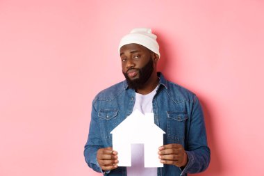 Emlak konsepti. Şüpheli siyah erkek model kağıt ev modelini tutuyor, şüpheli ve şüpheli görünüyor, daireyi arıyor, pembe arka planda duruyor.