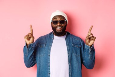 Mutlu sakallı Afro-Amerikalı adam anons yapıyor, parmaklarını havaya kaldırıyor ve gülümsüyor, bereli ve pembe arka planda güneş gözlüğü takıyor.