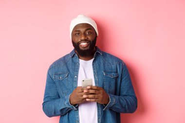 Yakışıklı Afro-Amerikalı adam akıllı telefon kullanıyor, kameraya gülümsüyor, pembe arka planda duruyor.