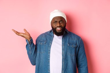 Sinirli ve kafası karışmış siyah adam tartışıyor, elini kaldırıyor ve kameraya şaşkın şaşkın bakıyor, ne olduğunu anlayamıyor, pembe arka planda duruyor.