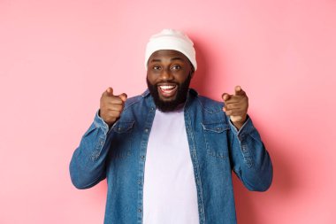 Mutlu Afro-Amerikalı hippi çocuk kamerayı işaret ediyor, sana ihtiyacı var, gülümsüyor ve şirkete davette bulunuyor, pembe arka planda duruyor.