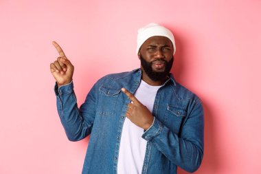 Kafası karışmış ve hayal kırıklığına uğramış Afro-Amerikan bir adam yüzünü ekşitmiş, sol üst köşedeki afişi işaret ediyor, iğrenç yüzüyle reklamını yapıyor, pembe arka planda duruyor.