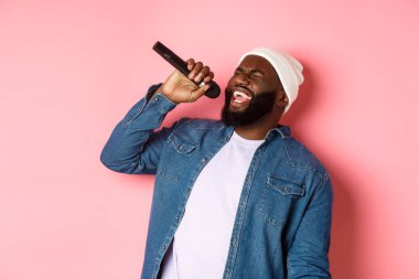 Bereli ve kot gömlekli yakışıklı siyah adam karaoke söylüyor, mikrofon tutuyor, pembe arka planda duruyor.