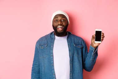 Bereli ve kot gömlekli yakışıklı hippi gülümsüyor, mutlu yüzlü cep telefonu ekranını gösteriyor, uygulama öneriyor, pembe arka planda duruyor.