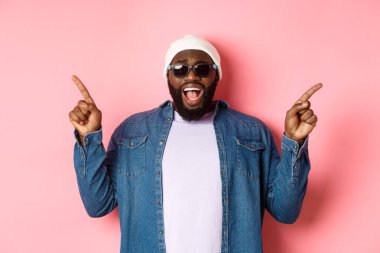 Harika havalı siyahi adam iki farklı şekil gösteriyor, parmaklarını yana doğru gösteriyor ve kameraya bakıyor, etkilenmiş, güneş gözlüğü takıyor, pembe arka planda duruyor.