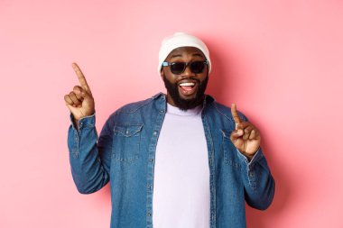 Mutlu sakallı Afro-Amerikalı adam anons yapıyor, sol üst köşeyi işaret ediyor ve gülümsüyor, bereyle ve pembe arka planda güneş gözlüğü takıyor.