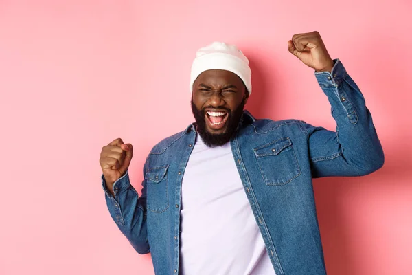 Szerencsés afro-amerikai srác nyert díjat, felemelte a kezét a diadal gesztus, sikoly öröm igen, ünnepli győzelem, rózsaszín háttér — Stock Fotó