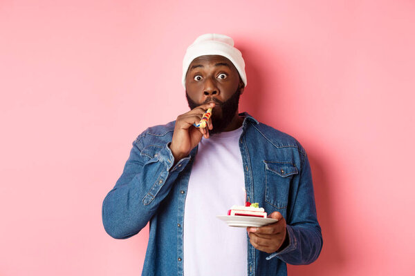 Happy Black hipster celebrating birthday, blowing party whistle, holding bday cake with candle, standing over pink background