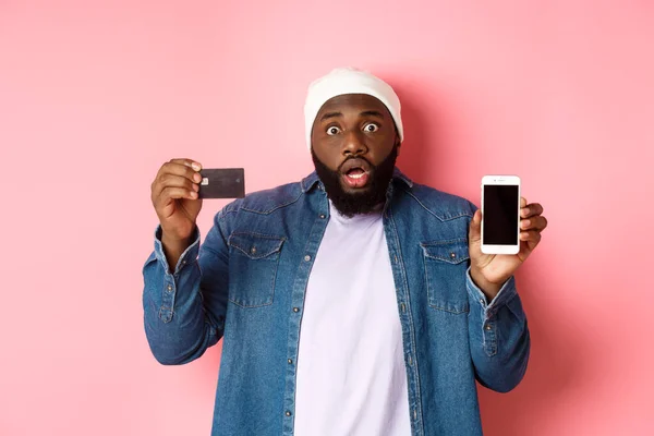 Online shopping. Shocked and concerned Black man staring at camera, showing mobile phone screen and credit card, standing over pink background