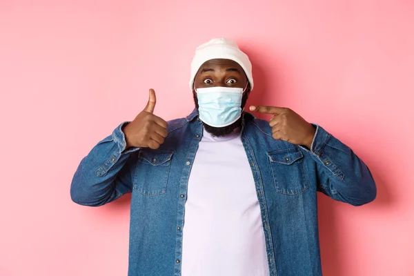 Coronavirus, τρόπος ζωής και παγκόσμια πανδημία έννοια. Νεαρός αφροαμερικανός που δείχνει τη μάσκα προσώπου και δείχνει τους αντίχειρες προς τα πάνω, προστατεύει τον εαυτό του από το ροζ φόντο. — Φωτογραφία Αρχείου