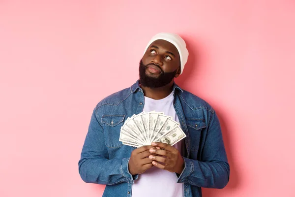 Homme afro-américain rêveur songeant à faire du shopping, tenant des dollars d'argent et regardant coin supérieur gauche, debout sur fond rose — Photo