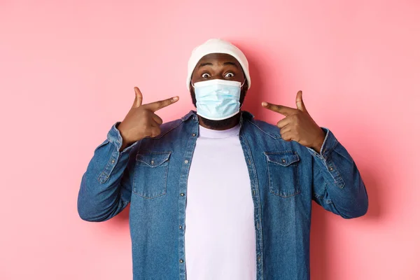 Coronavirus, τρόπος ζωής και την κοινωνική έννοια αποστασιοποίηση. Χαρούμενος μαύρος άντρας με σκούφο να σημαδεύει τη μάσκα του, χαμογελώντας στην κάμερα, να στέκεται πάνω από ροζ φόντο — Φωτογραφία Αρχείου