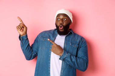 Şapkalı ve kot gömlekli etkileyici hippi siyahi adam sol üst köşedeki logoya bakıyor ve işaret ediyor, şaşkın şaşkın bakıyor, pembe arka planda duruyor.