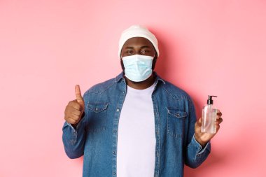 Covid-19, yaşam tarzı ve tecrit konsepti. Yüz maskesi ve bereli yakışıklı Afro-Amerikan hippi, el dezenfektanı ve baş parmak temizleyicisi, antiseptik