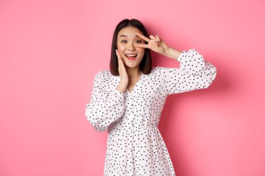 Güzel ve kadınsı Asyalı kadın kawaii işaretini gözlerinde gösteriyor ve kameraya hayret içinde bakıyor, pembe arka planda elbiseler içinde duruyor.