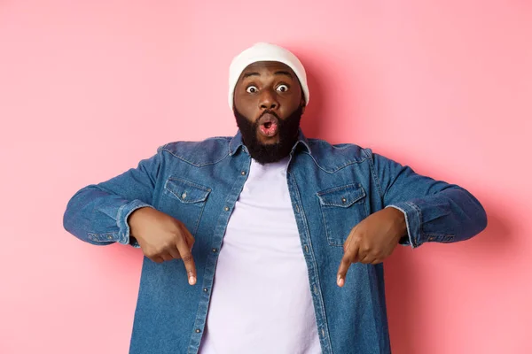 Verbazingwekkend en onder de indruk Afrikaans-Amerikaanse man tonen reclame, staren naar camera gelukkig en wijzen vingers naar beneden, roze achtergrond — Stockfoto