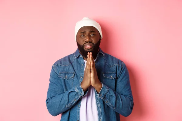 Upset african-american man asking for help, holding hands in pray, staring at camera sad and pleading, standing over pink background