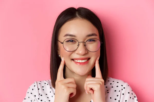 Concept beauté. Headshot de fille asiatique adorable dans des lunettes à la mode souriant, piquer les joues et montrant des fossettes mignonnes, debout sur fond rose — Photo