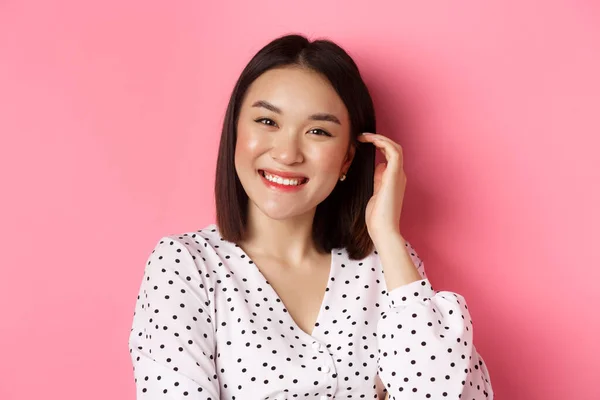 Primo piano di bella donna asiatica sorridente felice, toccante nuovo taglio di capelli, in piedi su sfondo rosa — Foto Stock