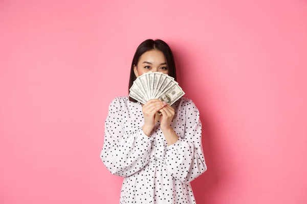 Концепция покупок. Милая азиатка, прячущая лицо за деньгами, подглядывающая в камеру, стоящая на розовом фоне — стоковое фото