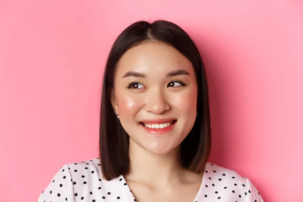 Conceito de beleza e estilo de vida. Headshot de mulher asiática bonita sonhadora olhando para a esquerda no espaço cópia, sorrindo feliz, de pé sobre fundo rosa — Fotografia de Stock