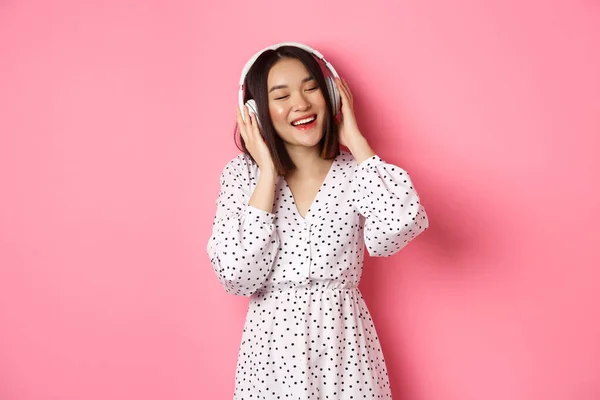 Romanttinen aasialainen nainen hymyilee onnellinen, kuuntelee musiikkia kuulokkeet ja tanssi, seisoo trendikäs mekko vaaleanpunainen tausta — kuvapankkivalokuva